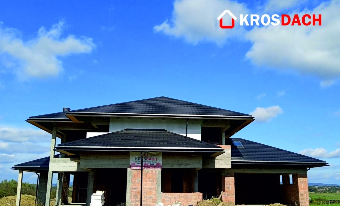 Dom w trakcie budowy z nowym, czarnym dachem z dachówki ceramicznej, widoczne rynny, na elewacji baner reklamowy firmy dekarskiej Krosdach, błękitne niebo z chmurami w tle.