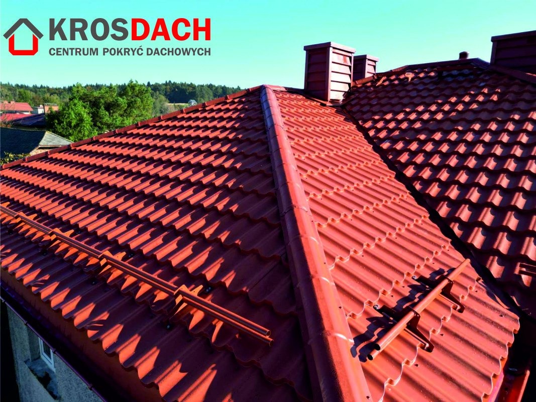 Czerwona dachówka modułowa na skośnym dachu z widocznymi kominami i logo firmy Krosdach w tle.