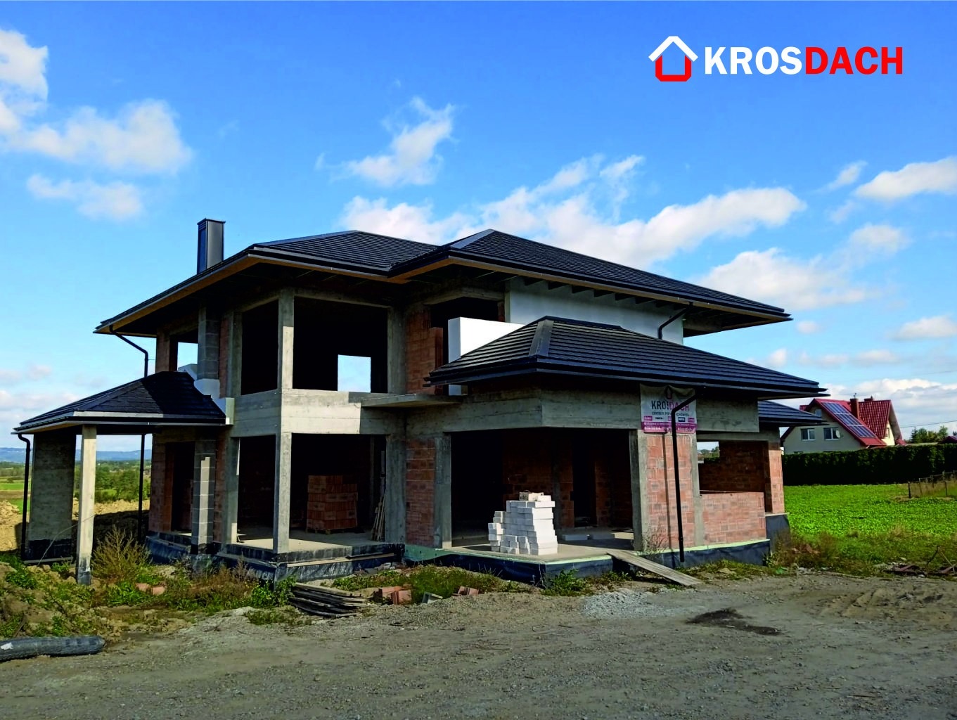 Niedokończony dom jednorodzinny z częściowo położonym, czarnym dachem, widoczne rynny i komin, logo firmy na górze kadru, w tle błękitne niebo z chmurami.