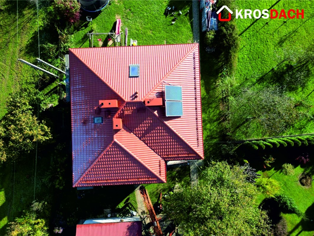 Widok z góry na dom z nowym, czerwonym dachem z dachówki ceramicznej, z oknem dachowym i panelami solarnymi, otoczony zielonym trawnikiem i drzewami. Widoczny znak firmowy Krosdach.