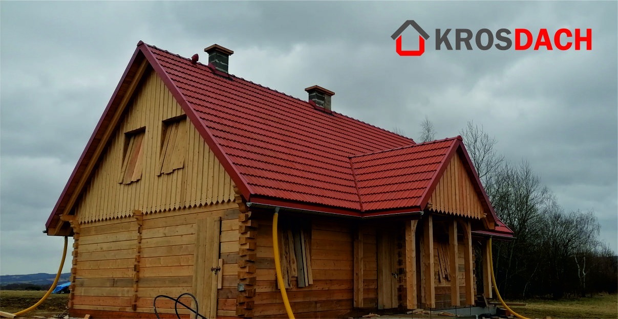 Nowo wybudowany drewniany dom z czerwoną dachówką, widoczne rynny i dwa kominy, na dachu logo firmy dekarskiej.