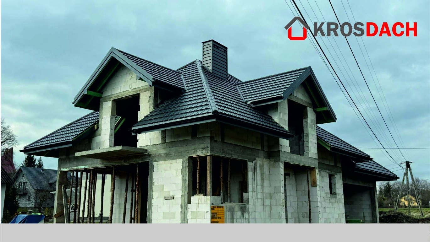 Dom w trakcie budowy z zamontowanym, czarnym dachem z dachówki ceramicznej i kominem, widoczne bloczki betonowe, szalunki i logo producenta na górze.