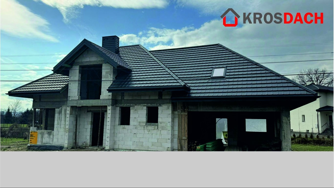 Dom w trakcie budowy z nowym, ciemnoszarym dachem z dachówki, widoczne okno dachowe i komin, ściany z białego bloczka, w tle błękitne niebo z chmurami i logo firmy Krosdach w prawym górnym rogu.