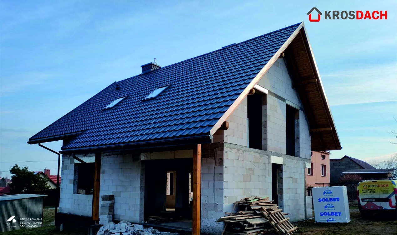 Niedokończony dom jednorodzinny z ciemnym, nowym pokryciem dachowym i oknami dachowymi, widoczne pustaki, brak okien i drzwi, materiały budowlane Solbet obok.