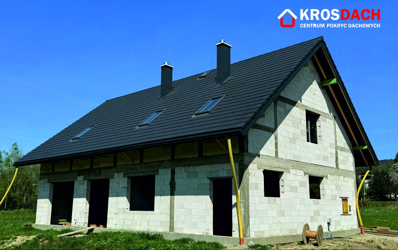 Dom w trakcie budowy z nowym, ciemnoszarym pokryciem dachowym, widoczne okna dachowe i kominy, ściany z białych bloczków, żółte rury spustowe i logo producenta na górze kadru.