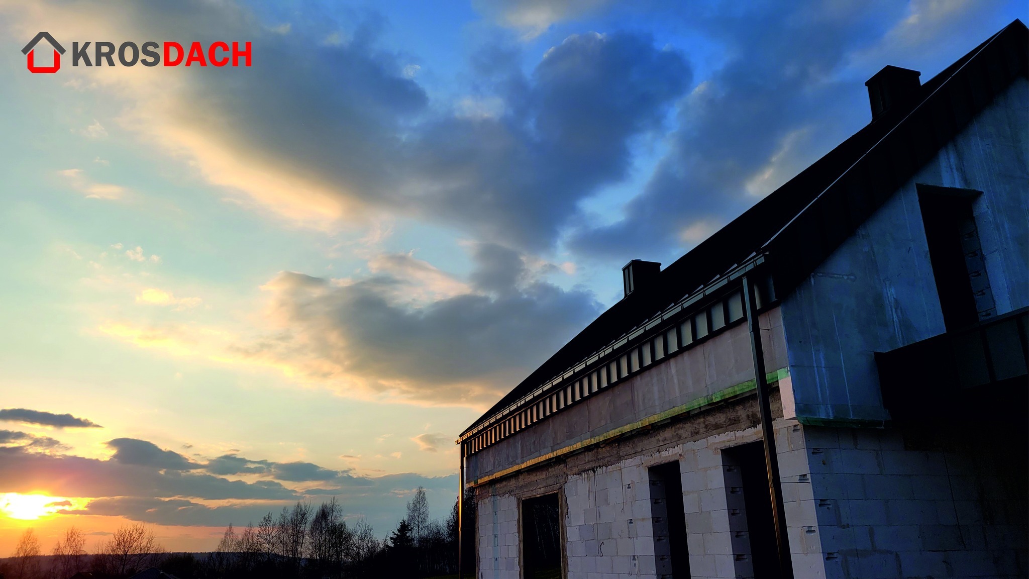 Niedokończony budynek z zamontowanym dachem i rynnami, widok z dołu na tle zachodzącego słońca i logo firmy Krosdach w lewym górnym rogu.