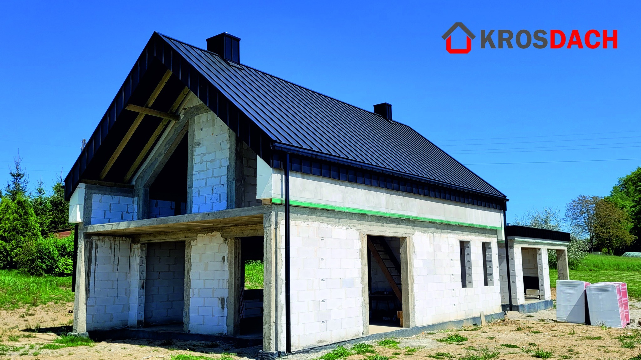 Dom w trakcie budowy z zamontowanym, czarnym dachem z blachy na rąbek stojący, widoczne rynny w kolorze dachu, ściany z białego bloczka, logo firmy dekarskiej w prawym górnym rogu.