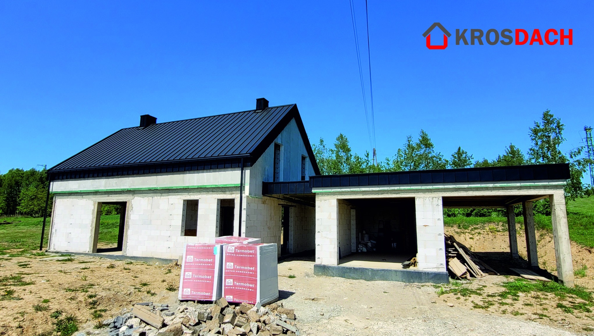 Dom w stanie surowym zamkniętym z ciemnym dachem z blachy na rąbek stojący, widoczne pakiety bloczków Termobet na pierwszym planie.