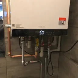 Biały piec gazowy Viessmann z wyświetlaczem pokazującym temperaturę 50 stopni, podłączony do instalacji z rur miedzianych i stalowych na tle płytek imitujących drewno.