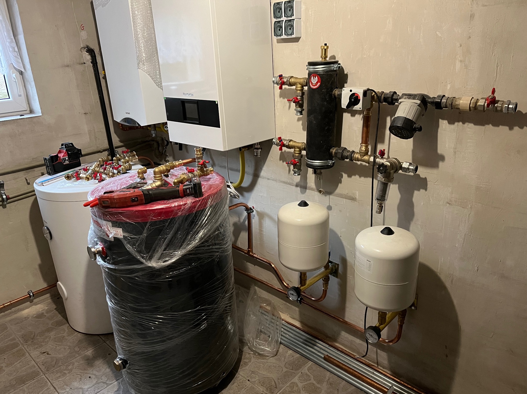 Nowoczesny system grzewczy z kotłem gazowym, zbiornikiem buforowym owiniętym folią, naczyniami wzbiorczymi i rozbudowaną instalacją hydrauliczną z zaworami, rurami miedzianymi i pompą obiegową.