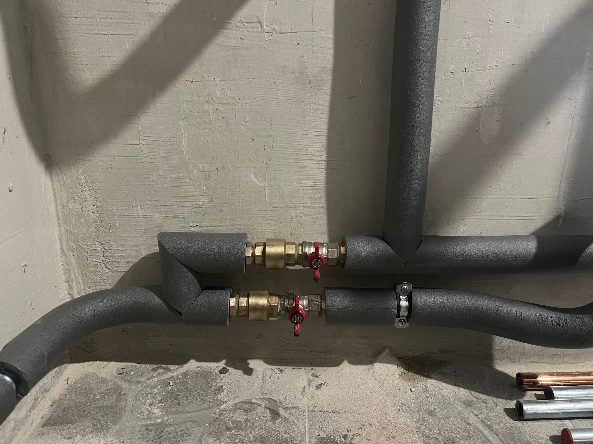 Dwie rury grzewcze z zaworami odcinającymi, zaizolowane szarą pianką, zamontowane przy surowej, jasnej ścianie. Fragmenty rur miedzianych i stalowych na podłodze.