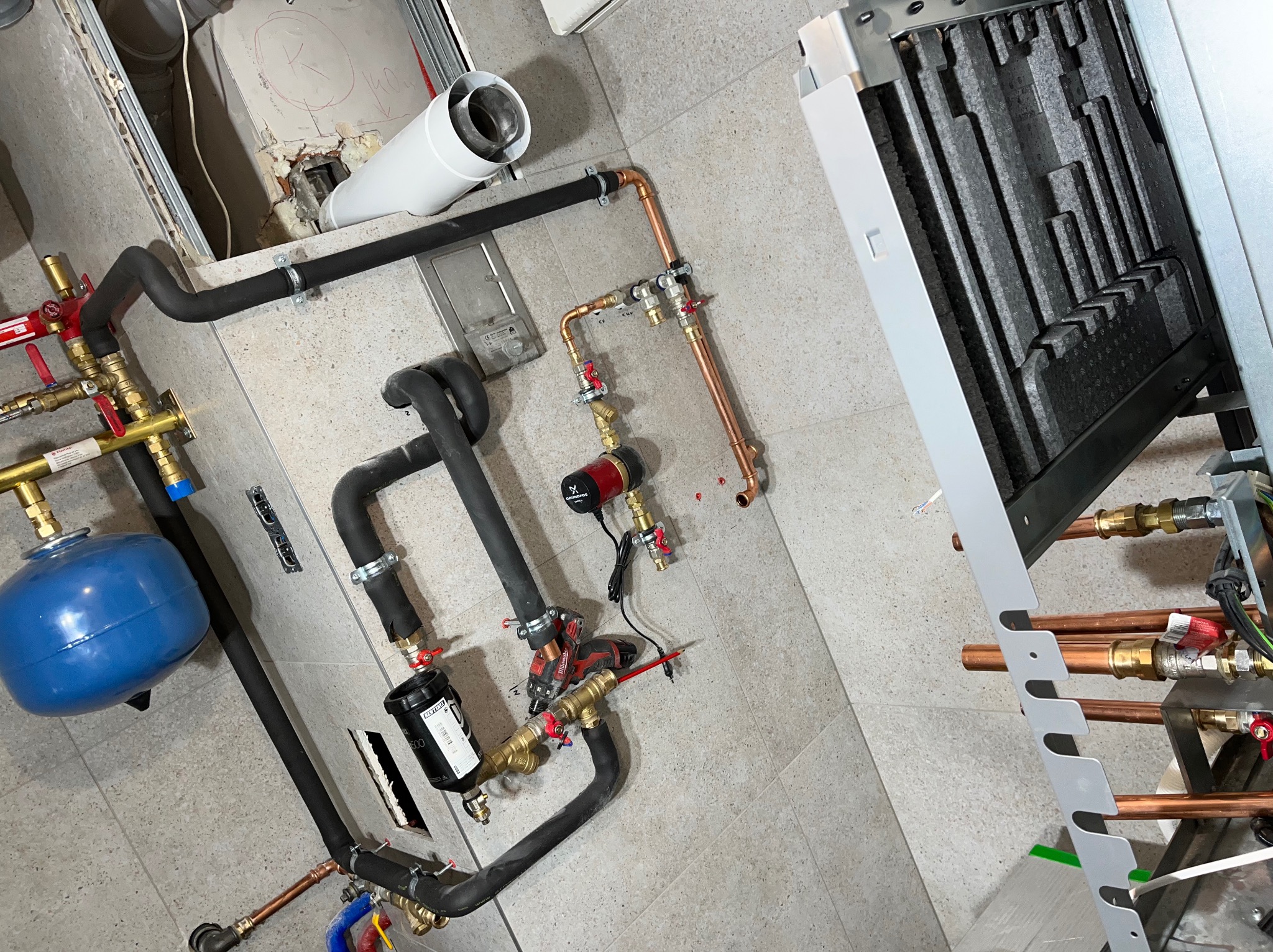 Widok z góry na instalację grzewczą w trakcie montażu, z widocznym kotłem gazowym, rurami miedzianymi, czarnymi rurami izolowanymi, niebieskim naczyniem przeponowym, pompą obiegową Grundfos...