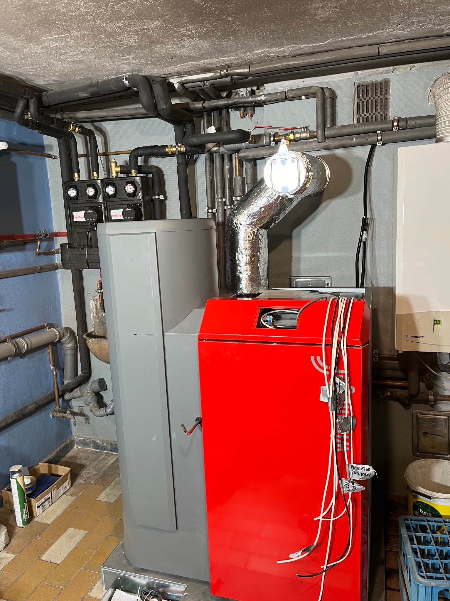 Nowoczesny piec z szarym korpusem i czerwonym panelem, z podłączonymi przewodami i aluminiową rurą odprowadzającą spaliny, widoczne liczne rury instalacyjne pod sufitem w piwnicy.