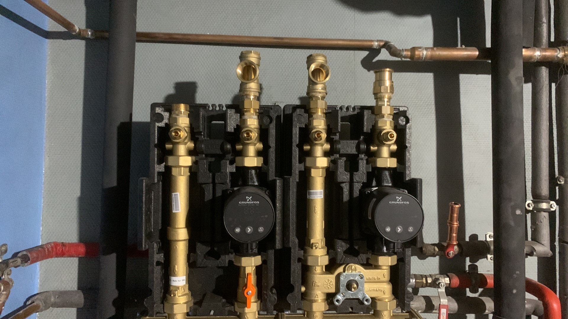 Rozdzielacz ogrzewania podłogowego z dwoma pompami obiegowymi Grundfos Alpha 2, widoczne mosiężne zawory i rury miedziane w szarej kotłowni.