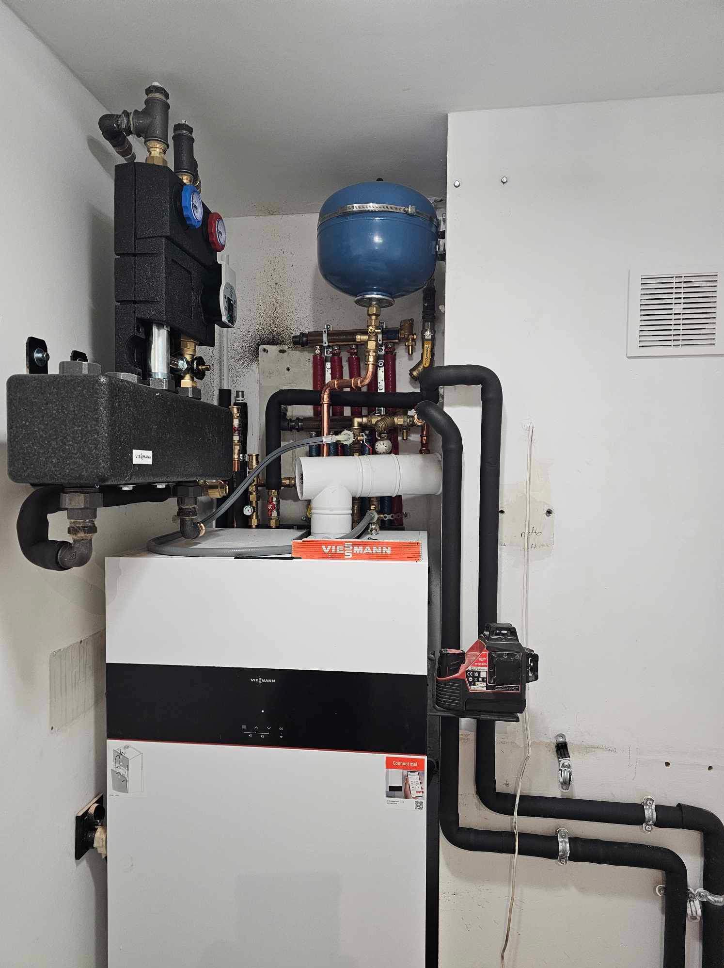 Nowoczesny piec gazowy Viessmann z czarnymi rurami instalacji centralnego ogrzewania, niebieskim zbiornikiem wyrównawczym i izolowanymi elementami armatury, zamontowany przy białej ścianie.