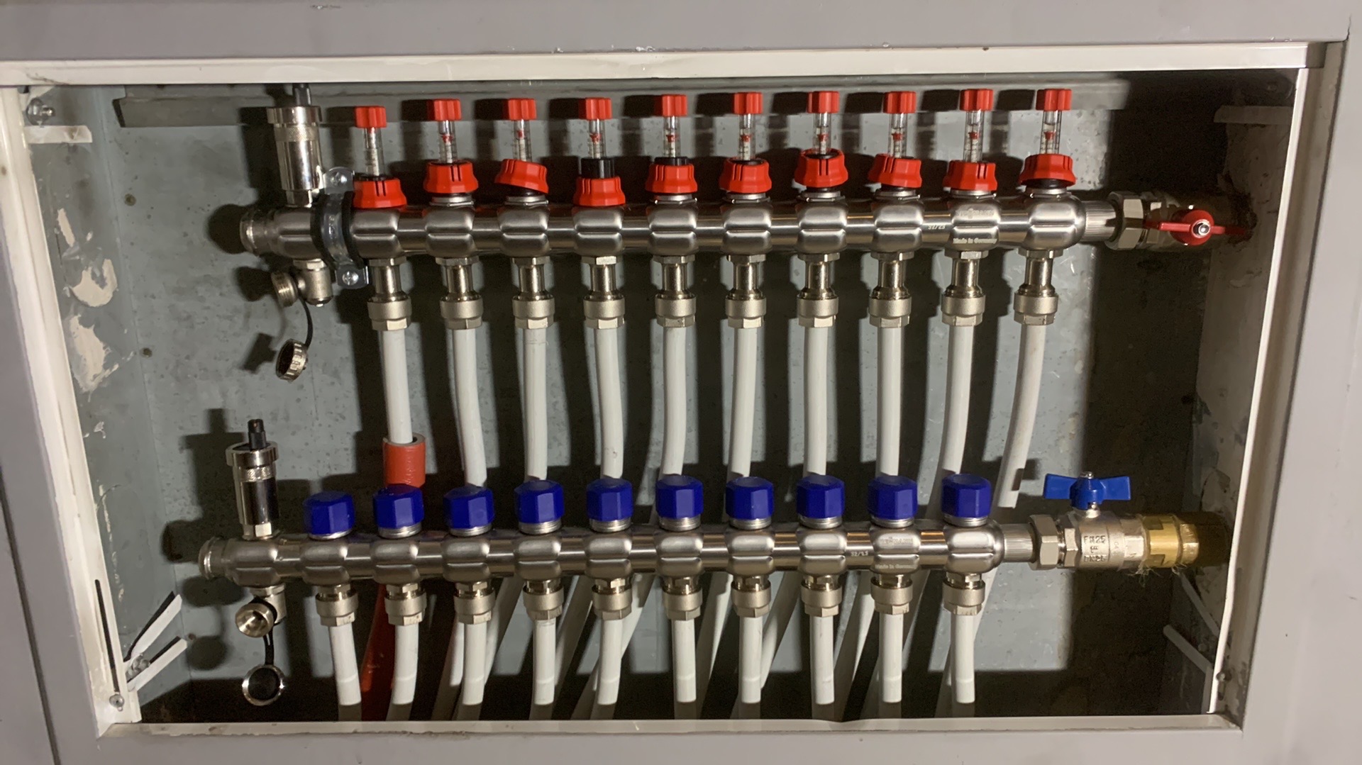 Rozdzielacz ogrzewania podłogowego w szarej szafce, widoczne zawory z czerwonymi i niebieskimi pokrętłami oraz białe rury.