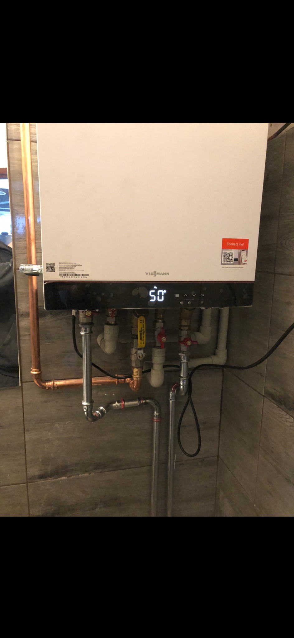 Biały piec gazowy Viessmann z wyświetlaczem pokazującym temperaturę 50 stopni, podłączony do instalacji z rur miedzianych i stalowych na tle płytek imitujących drewno.