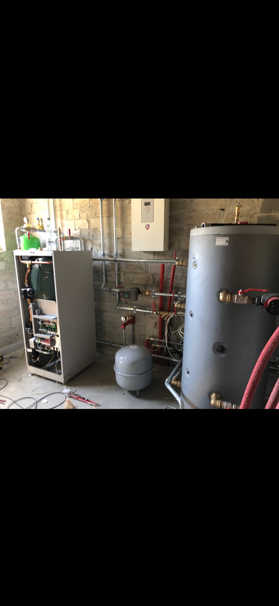 Wnętrze kotłowni w trakcie montażu, widoczny otwarty panel sterowania nowego pieca, zbiornik wyrównawczy ciśnienia i duży zasobnik ciepłej wody oraz liczne rury miedziane i plastikowe.