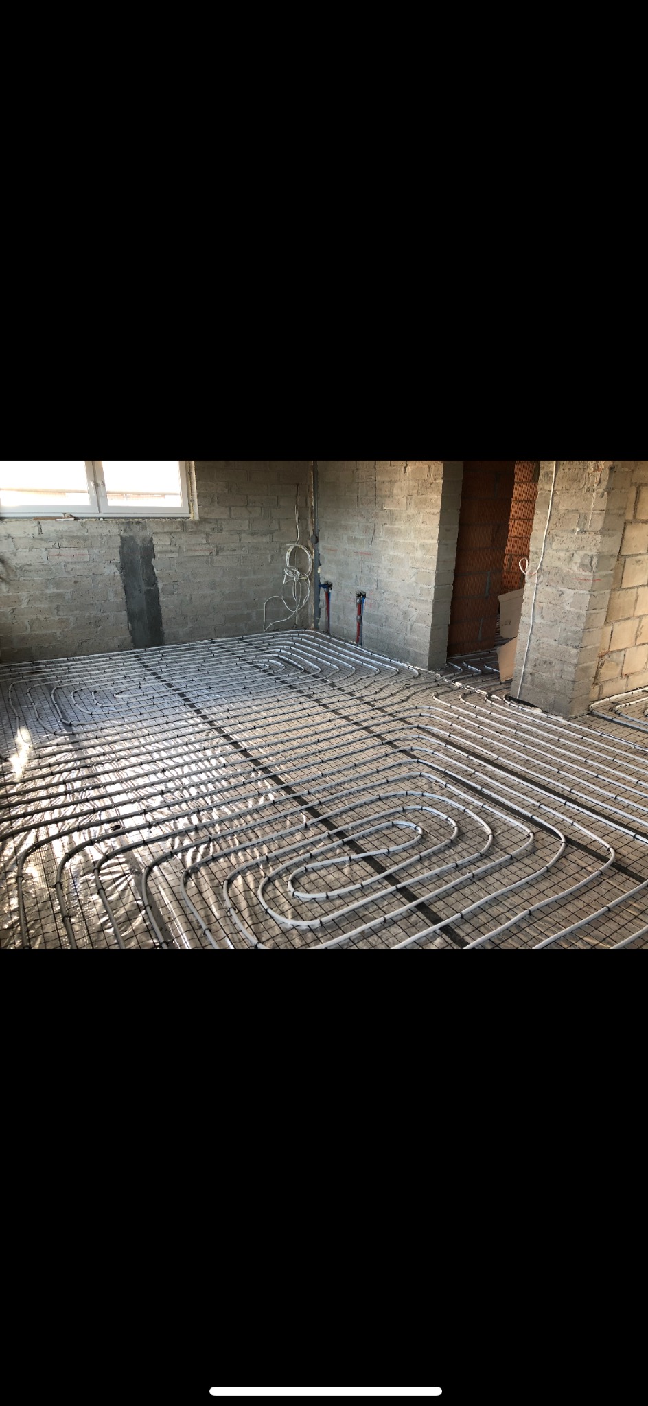 Montaż ogrzewania podłogowego: rury ułożone na folii izolacyjnej w surowym pomieszczeniu z ceglanymi ścianami, widoczne pionowe rury przyłączeniowe.