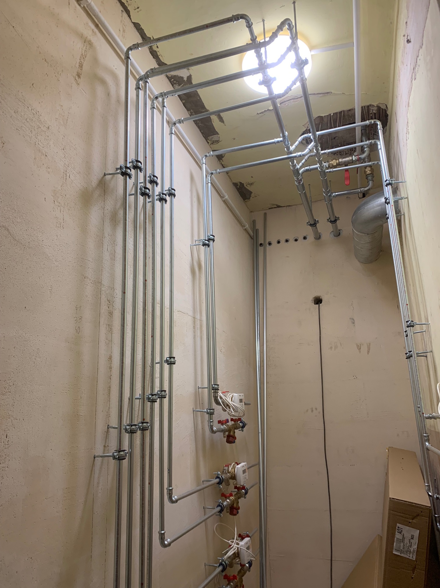 Szczegółowy widok instalacji rur grzewczych z zaworami i licznikami, zamocowanych na ścianie z surowym wykończeniem, widoczne elementy konstrukcji pod sufitem.