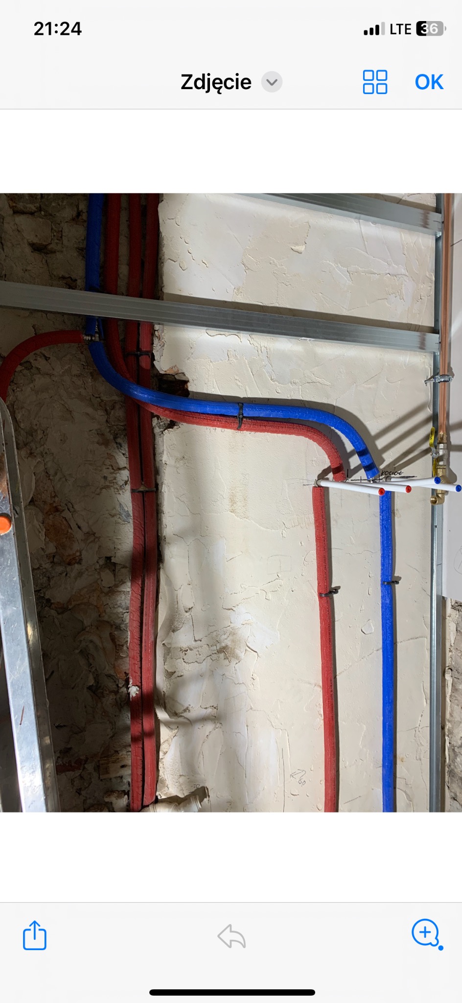 Wewnętrzna instalacja hydrauliczna z rurami w izolacji czerwonej i niebieskiej, przymocowanymi do ściany z surowym tynkiem, widoczne metalowe profile montażowe.