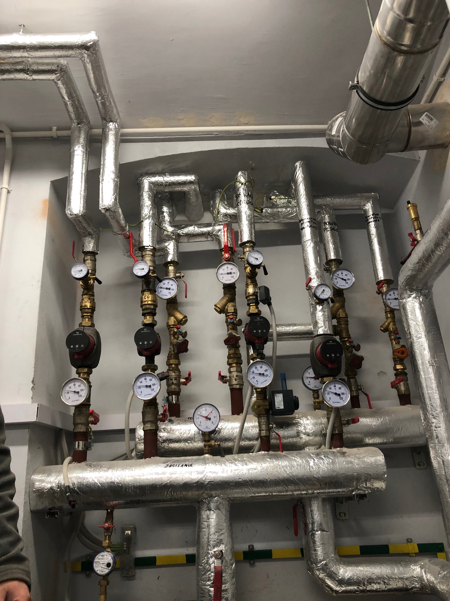 Rozbudowana instalacja grzewcza z izolowanymi rurami, licznymi zaworami, manometrami i pompami obiegowymi w kotłowni, widoczne napisy na rurach.