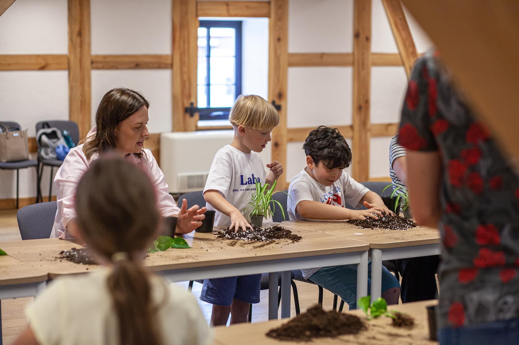 Dzieci z dorosłymi sadzą małe rośliny w doniczkach na warsztatach edukacyjnych, widoczne drewniane elementy konstrukcyjne budynku w tle.