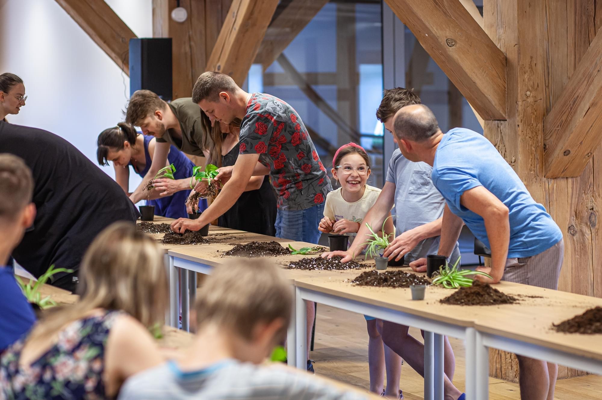 Grupa osób, w tym uśmiechnięta dziewczynka w okularach i opasce, uczestniczy w warsztatach sadzenia roślin doniczkowych na długim stole w jasnym pomieszczeniu z drewnianymi elementami konstrukcyjnymi