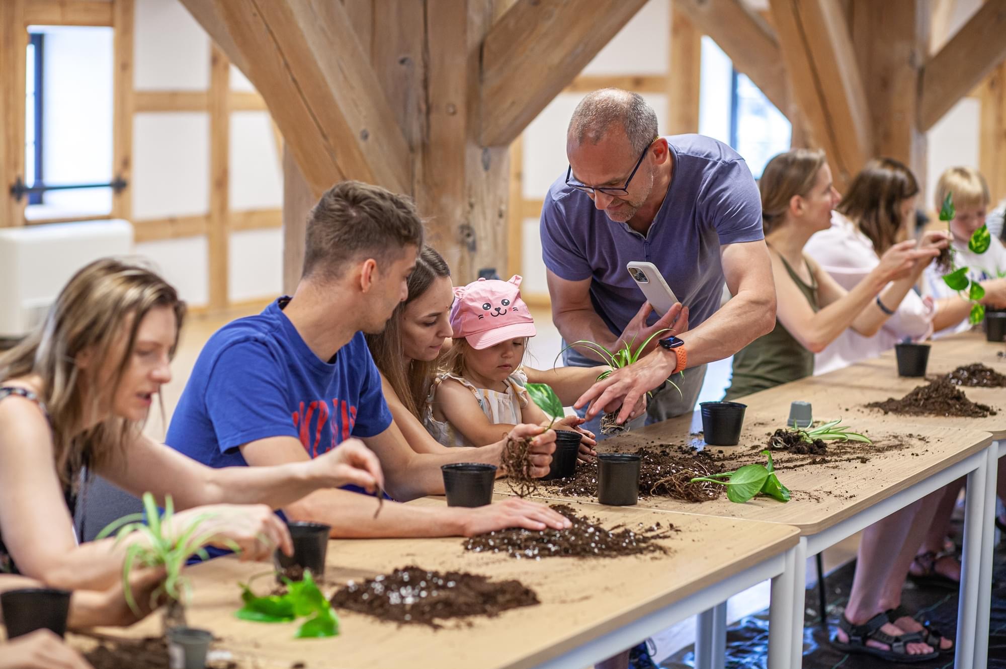 Grupa osób, w tym mała dziewczynka w różowej czapeczce, uczestniczy w warsztatach sadzenia roślin doniczkowych na długim drewnianym stole. Mężczyzna w okularach robi zdjęcie telefonem podczas...