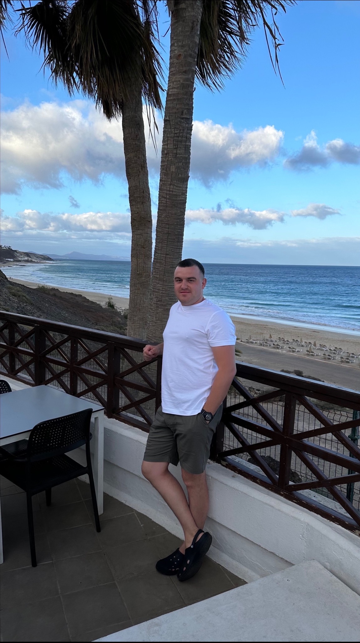 Mężczyzna oparty o balustradę balkonu z widokiem na plażę i ocean, w tle palmy i błękitne niebo z chmurami.