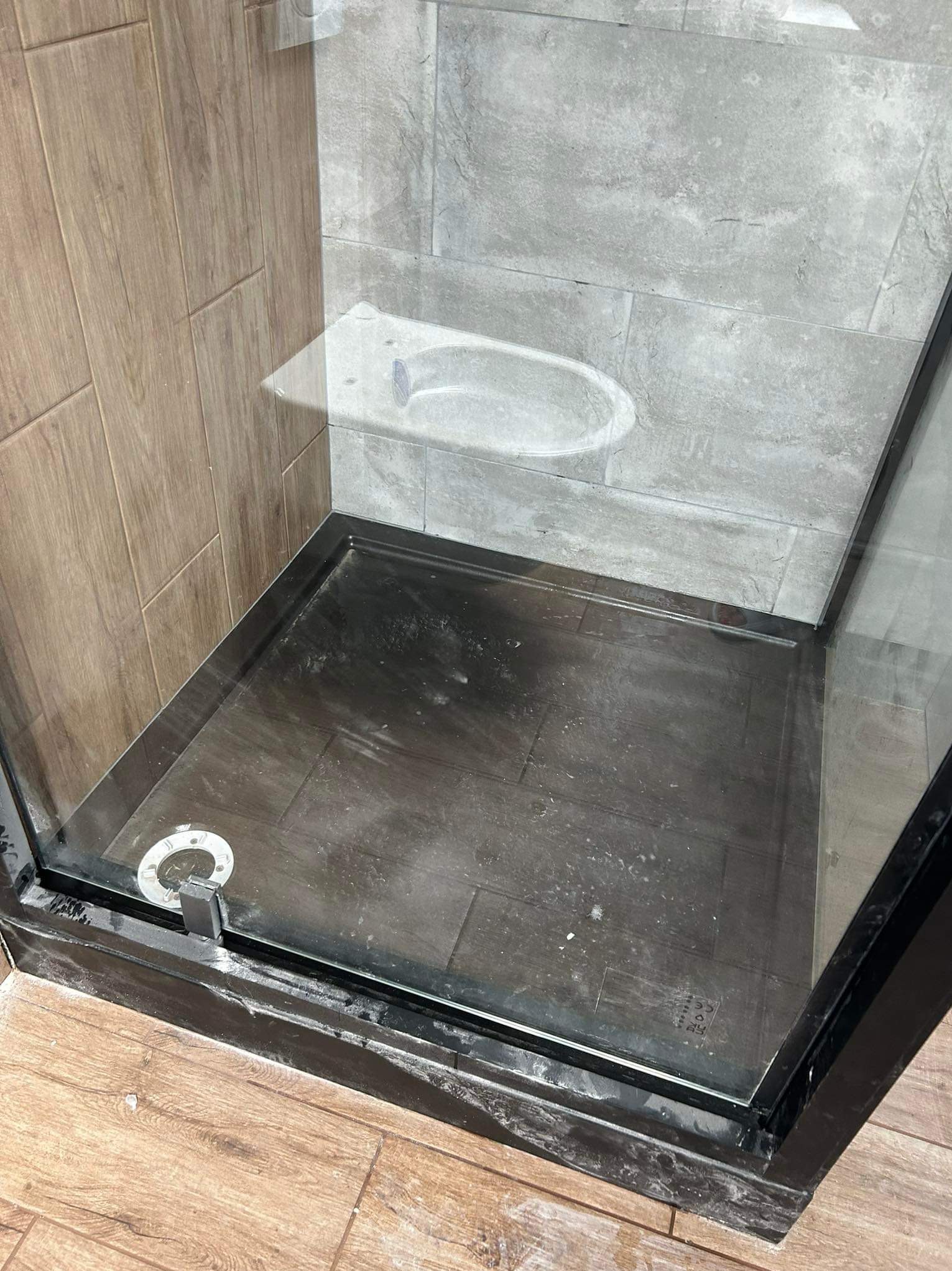 Brudny brodzik kabiny prysznicowej z ciemnymi płytkami, pozostałości po remoncie widoczne na fugach i elementach kabiny, fragment odpływu w rogu.