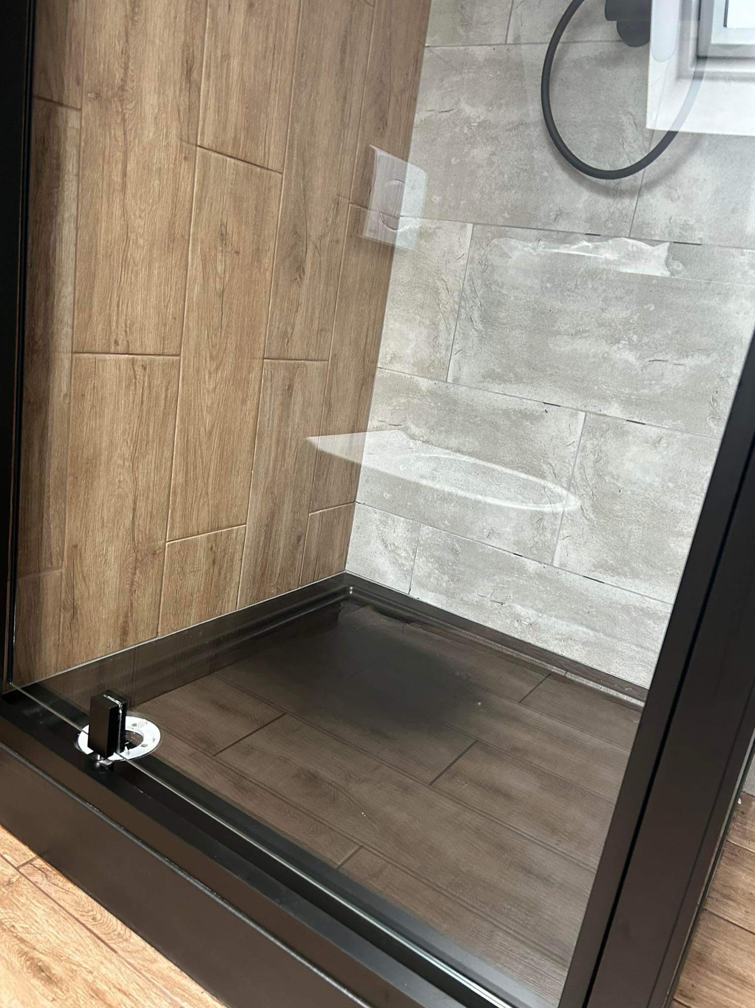 Nowoczesna kabina prysznicowa z ciemną ramą i imitacją drewna na ścianach i podłodze, widok z boku, szklana szyba z lekkimi zaciekami.