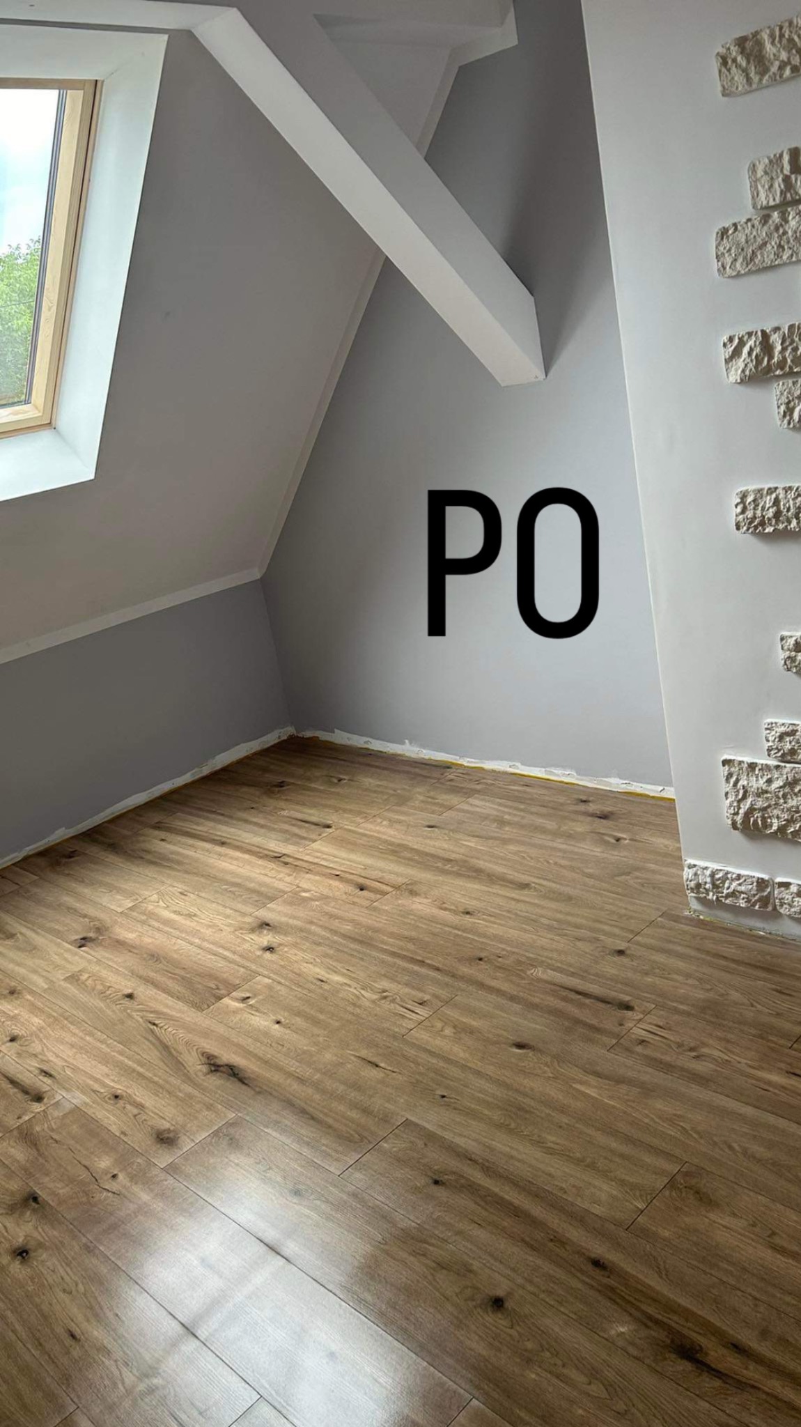 Poddasze po remoncie: nowa podłoga z paneli drewnopodobnych, ściany pomalowane na szaro, dekoracyjna ściana z białej cegły, okno dachowe wpuszczające światło.