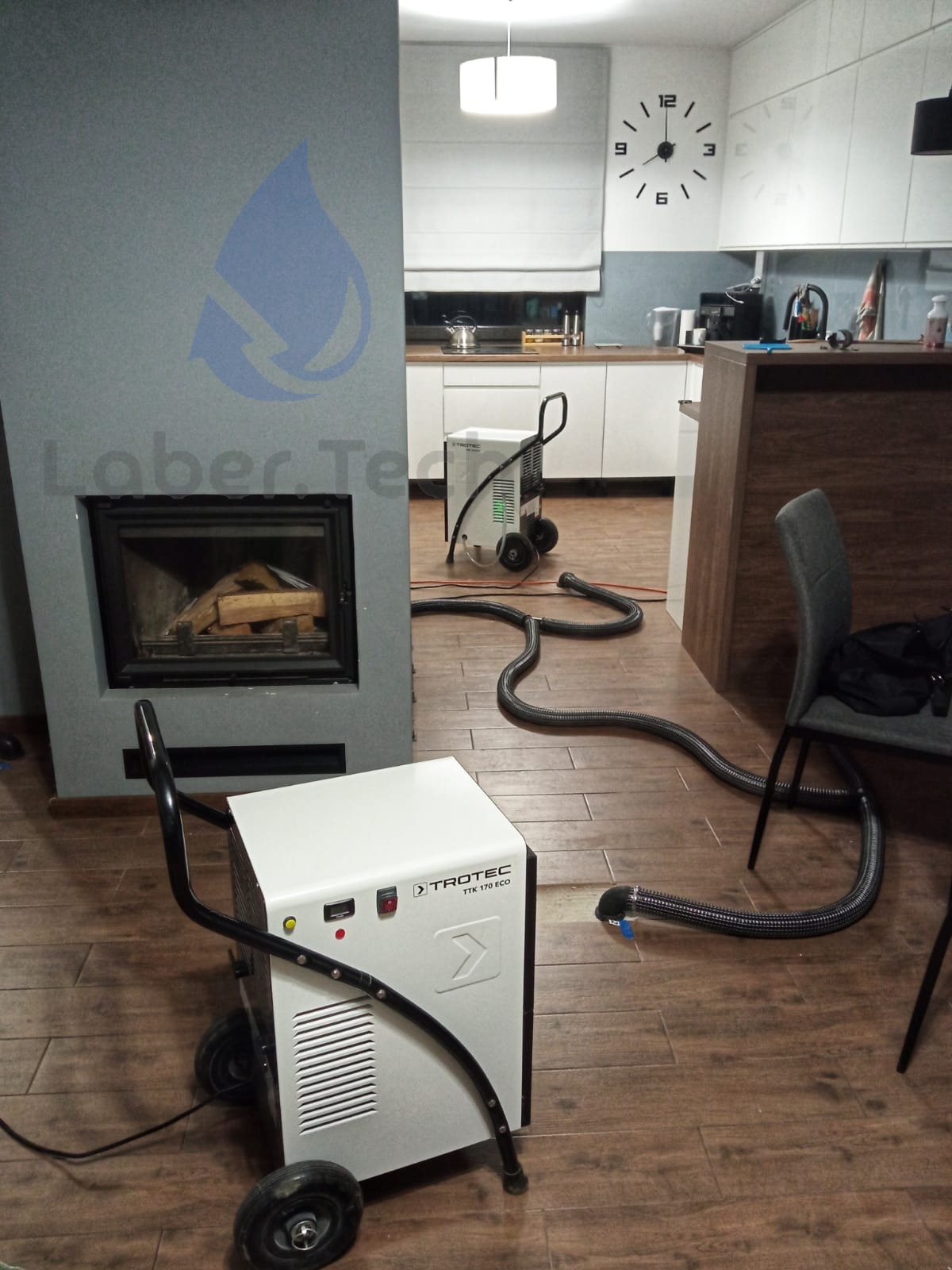 Dwa osuszacze powietrza Trotec TTK 170 ECO w pomieszczeniu z kominkiem i widoczną kuchnią w tle, w trakcie pracy z podłączonymi wężami odprowadzającymi wilgoć.