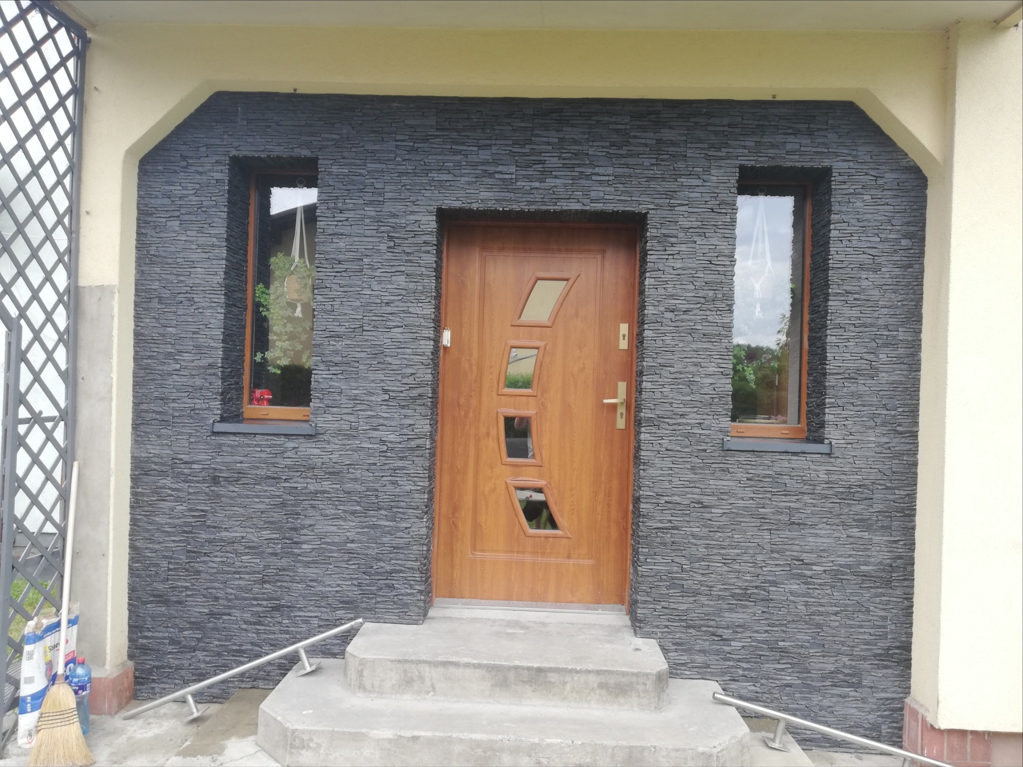 Elewacja budynku wykończona szarą kamienną okładziną z drewnianymi oknami i drzwiami z geometrycznymi przeszkleniami, widoczne schody wejściowe i fragment balustrady ze stali nierdzewnej oraz worki...