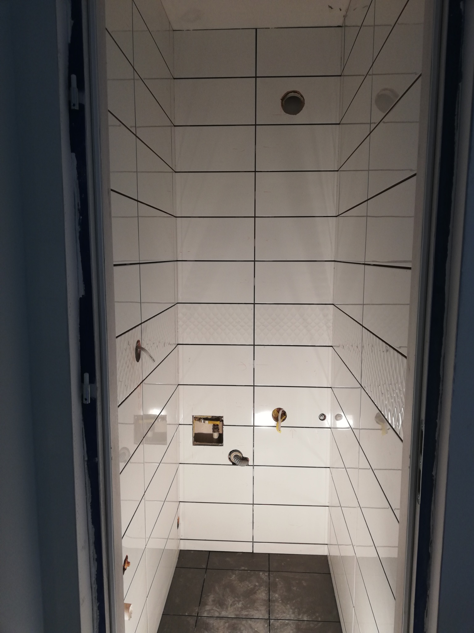 Wnętrze łazienki w trakcie prac wykończeniowych: białe płytki z ciemną fugą na ścianach i suficie, ciemna podłoga, widoczne instalacje hydrauliczne i elektryczne, otwarte otwory montażowe.