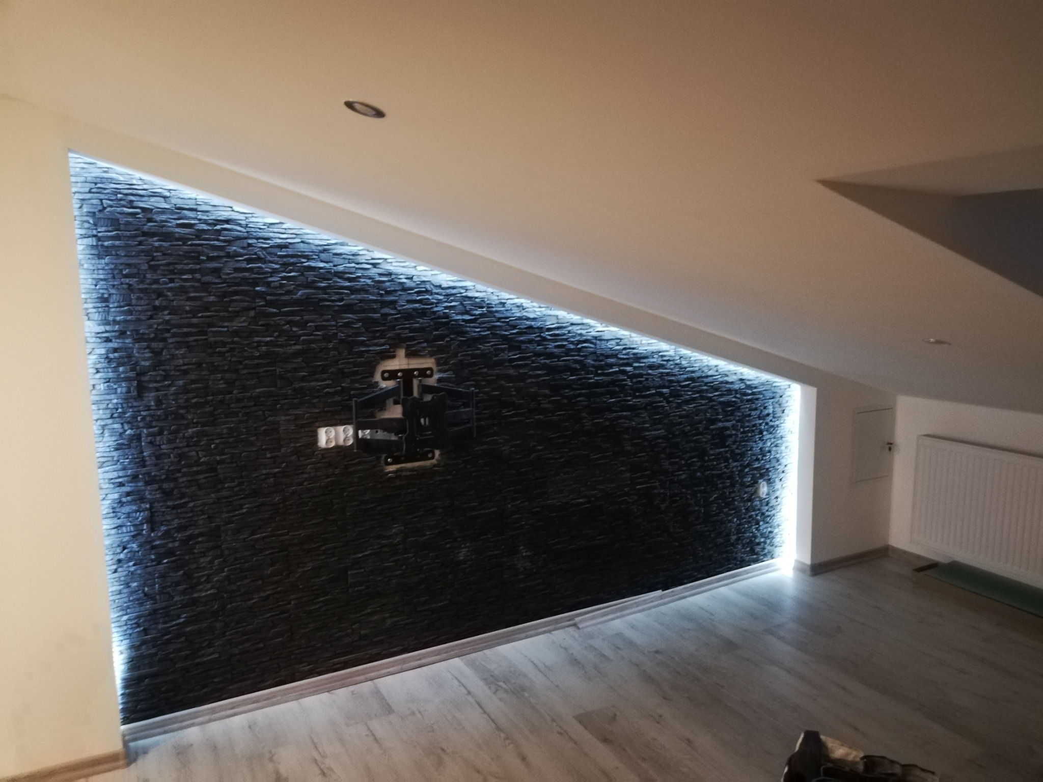 Ściana wykończona czarnym kamieniem dekoracyjnym z podświetleniem LED w skosie poddasza, z zamontowanym uchwytem do telewizora.