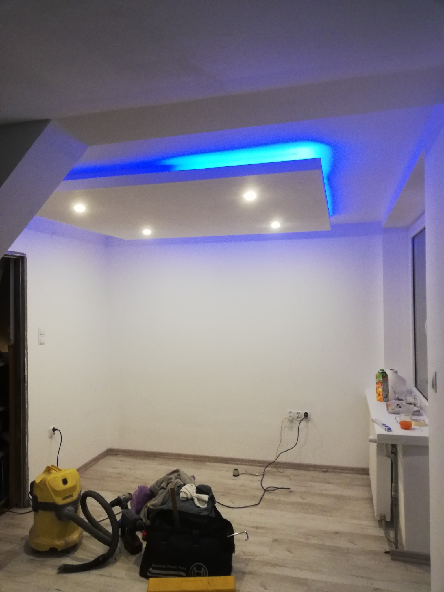 Pokój po remoncie z podwieszonym sufitem z oświetleniem LED w kolorze niebieskim i punktowymi lampami. Na podłodze widoczny odkurzacz przemysłowy i narzędzia.