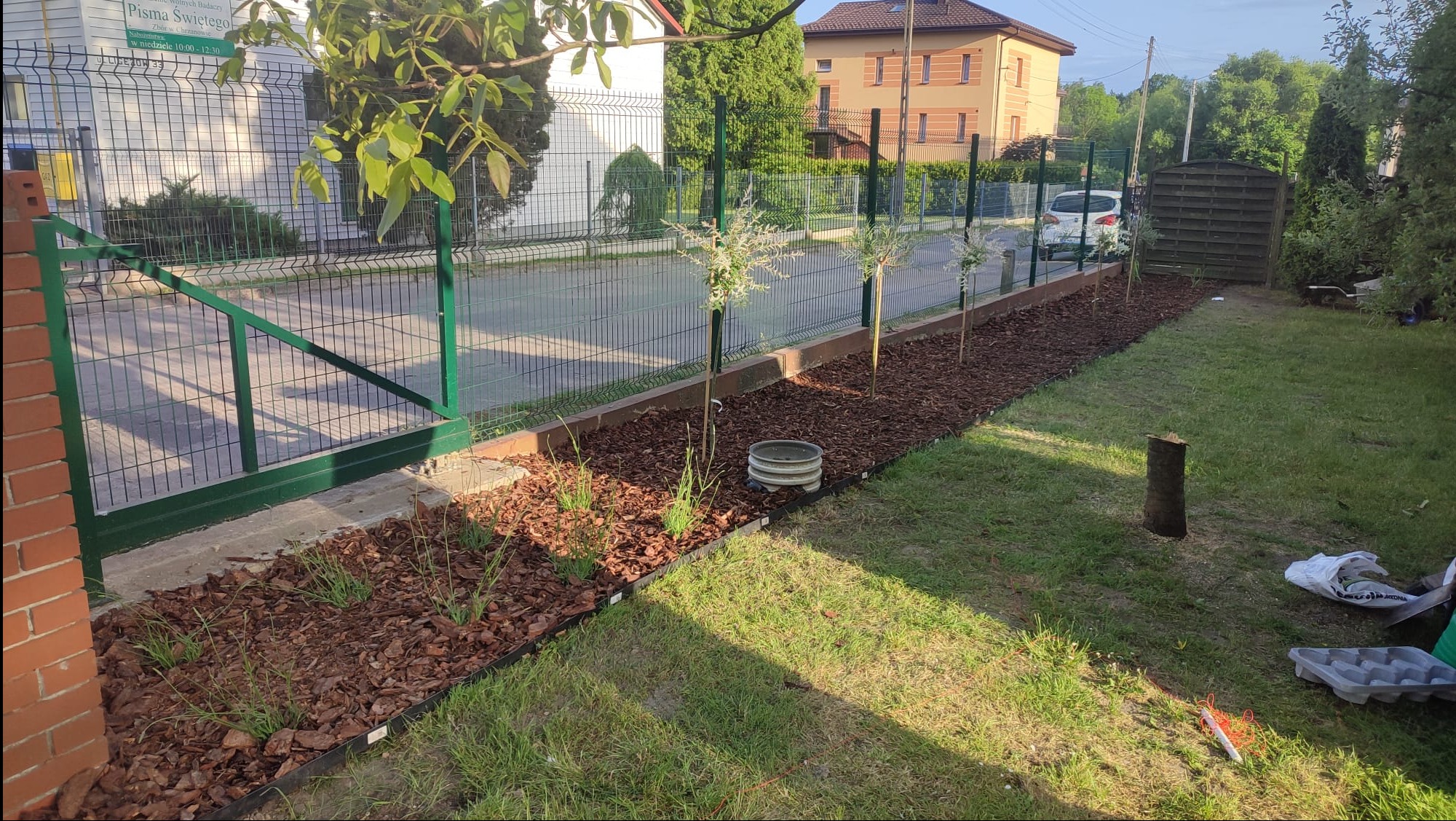 Świeżo założony trawnik z rabatą wyłożoną korą, wzdłuż ogrodzenia z metalowej siatki, z młodymi drzewkami i elementami dekoracyjnymi, widok na dom w tle.
