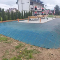 Niebieska siatka zabezpieczająca nasyp z ziemi, obok rosną tuje, a w tle budynek z parasolami. Przygotowanie terenu pod ogród.