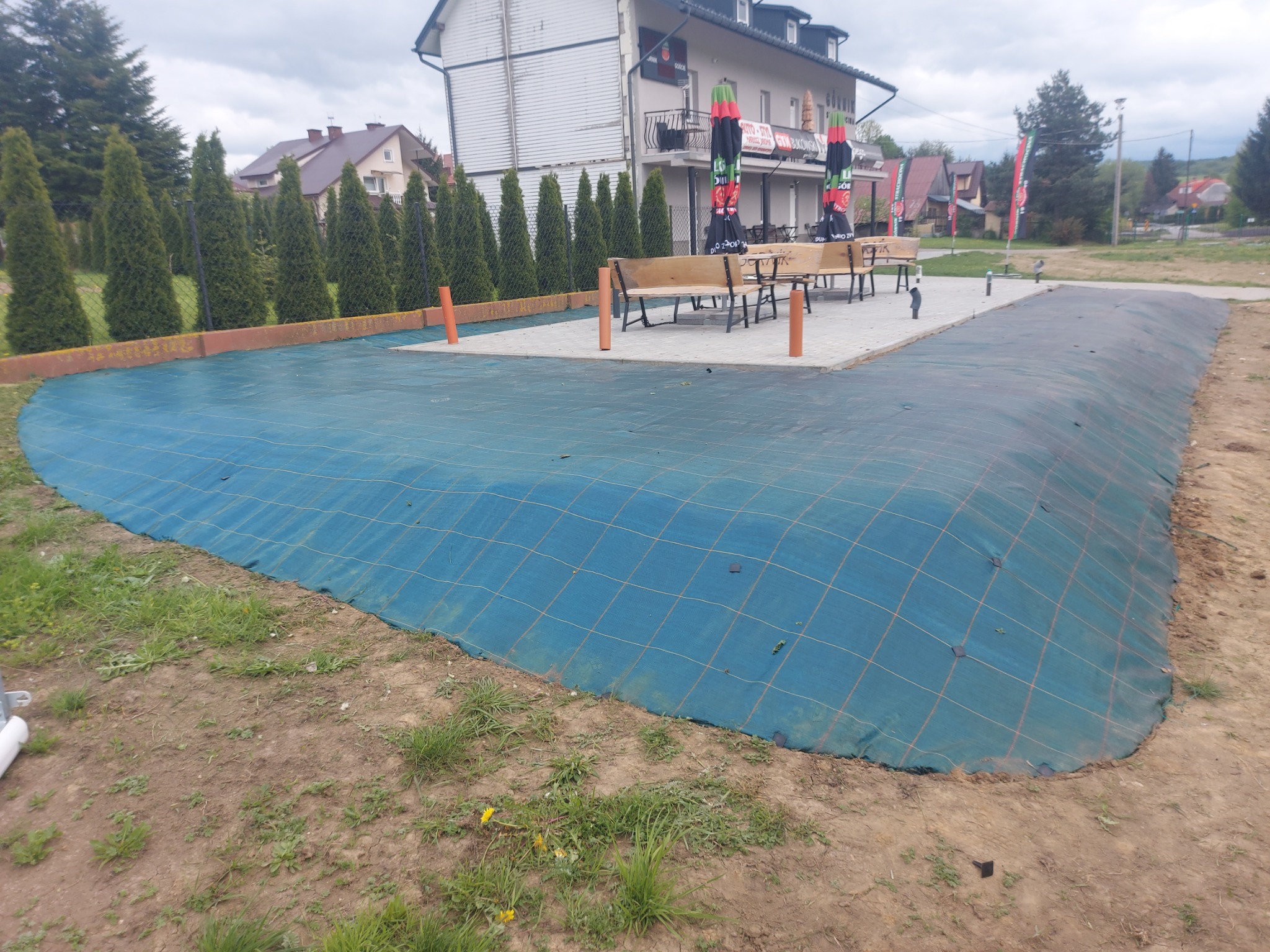 Niebieska siatka zabezpieczająca nasyp z ziemi, obok rosną tuje, a w tle budynek z parasolami. Przygotowanie terenu pod ogród.