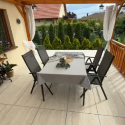 Zadaszony taras z beżową podłogą. Stół z krzesłami, dekoracje, radio na stoliku ze starej maszyny do szycia. W tle zielony ogród i białe zasłony.