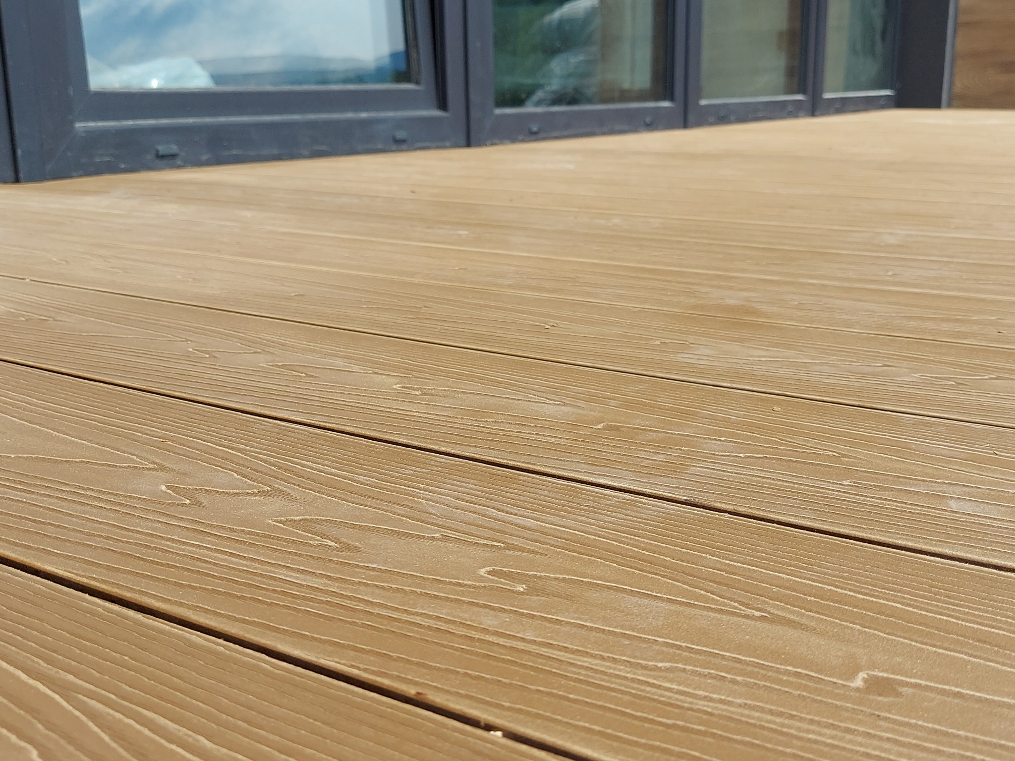 Jasnobrązowe deski tarasowe z widocznym rysunkiem słojów drewna, ułożone poziomo, z szarymi ramami okiennymi w tle.