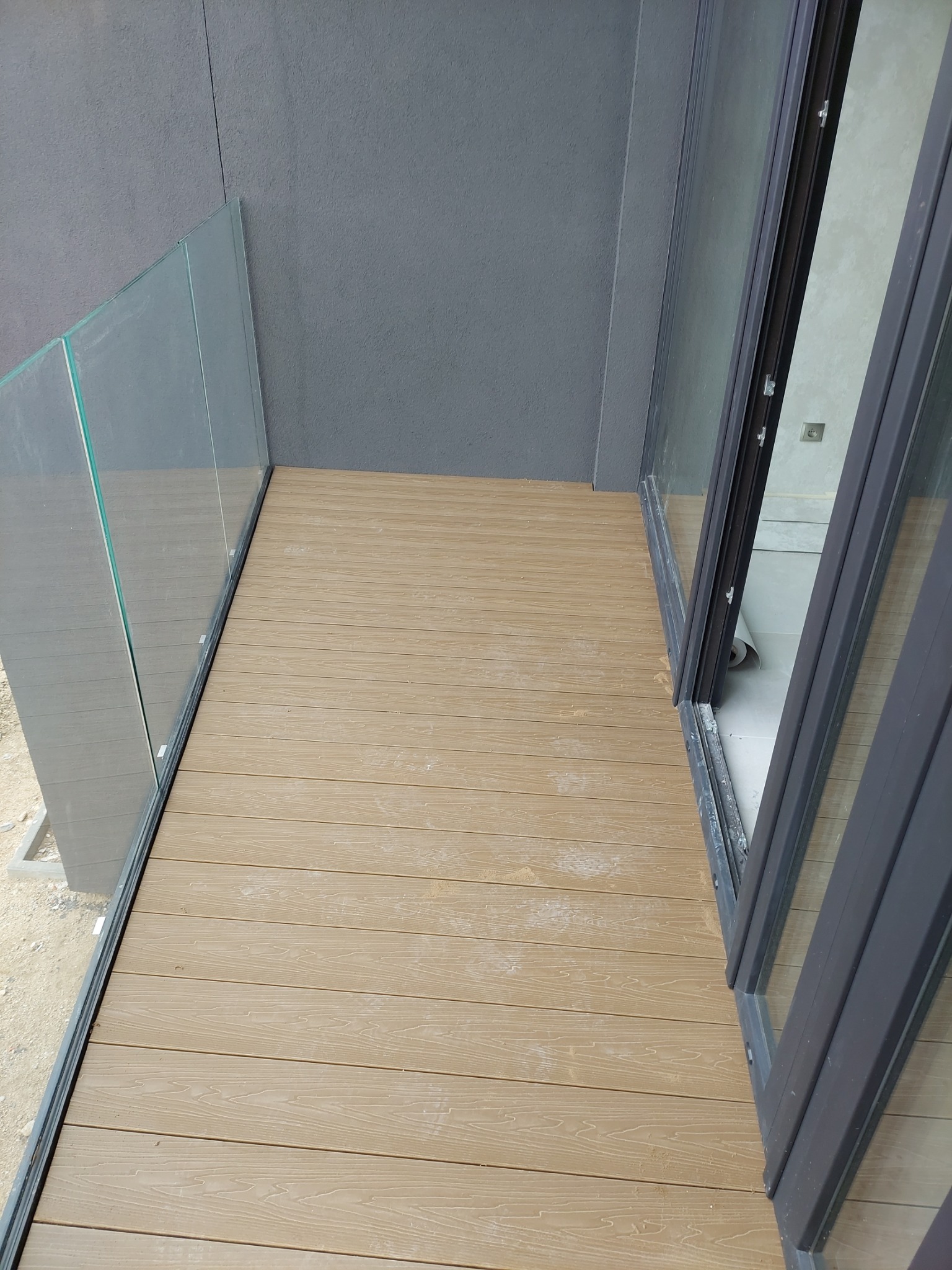 Nowoczesny balkon z deskami w odcieniach beżu i szklaną balustradą, widok z góry na świeżo położoną podłogę, w tle przesuwne drzwi balkonowe.
