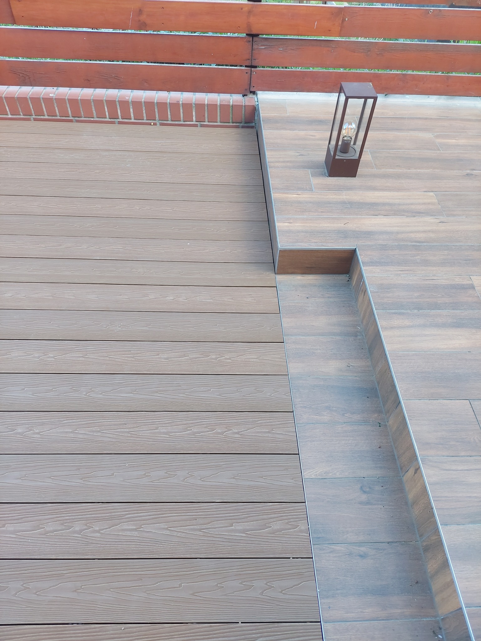 Nowoczesny taras z desek kompozytowych w odcieniach brązu, połączony z płytkami ceramicznymi imitującymi drewno, oświetlony lampą ogrodową