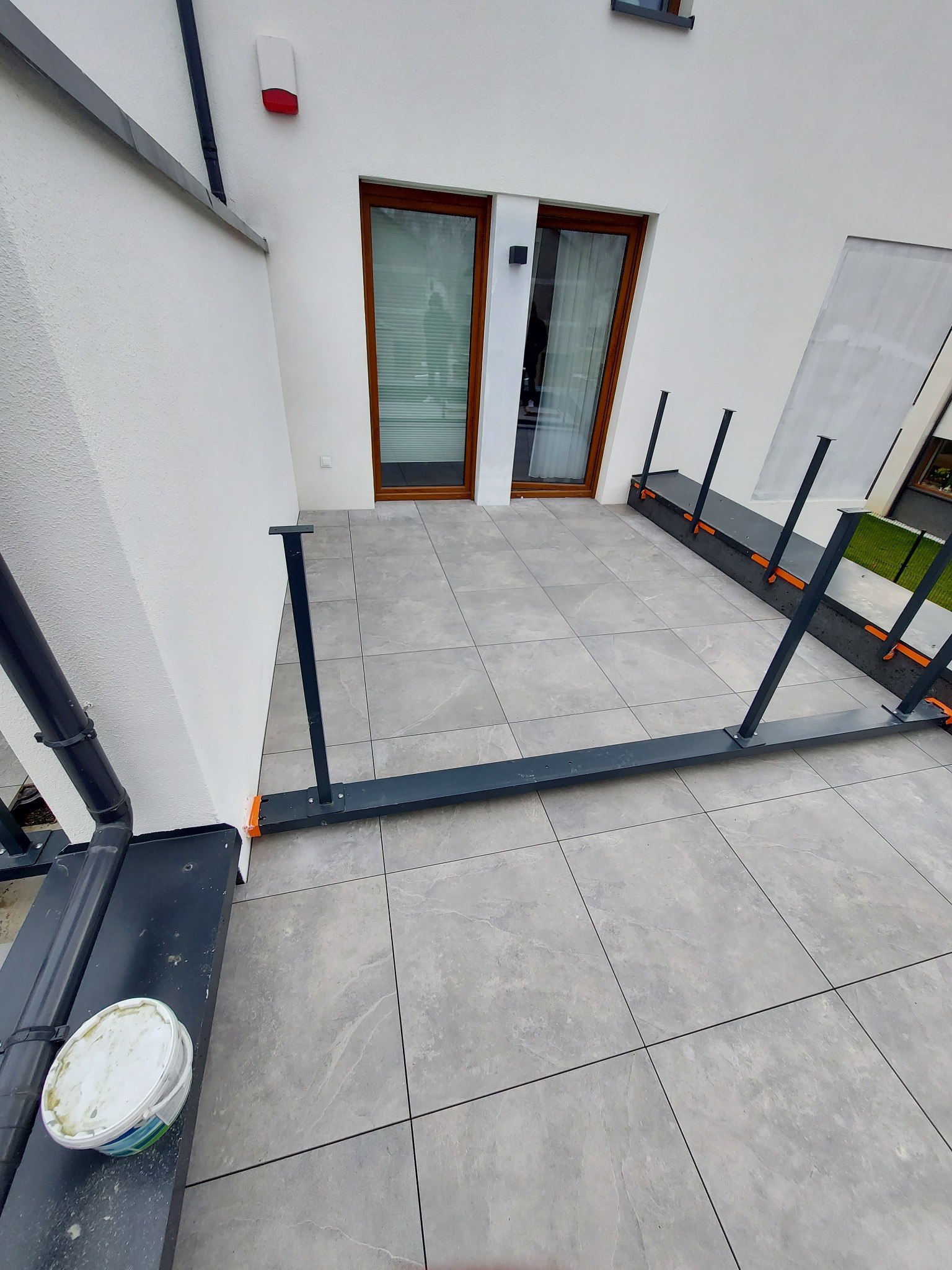 Montaż konstrukcji pod taras na szarych płytkach, widoczny kubełek z zaprawą i elementy mocujące do ściany budynku z oknami.