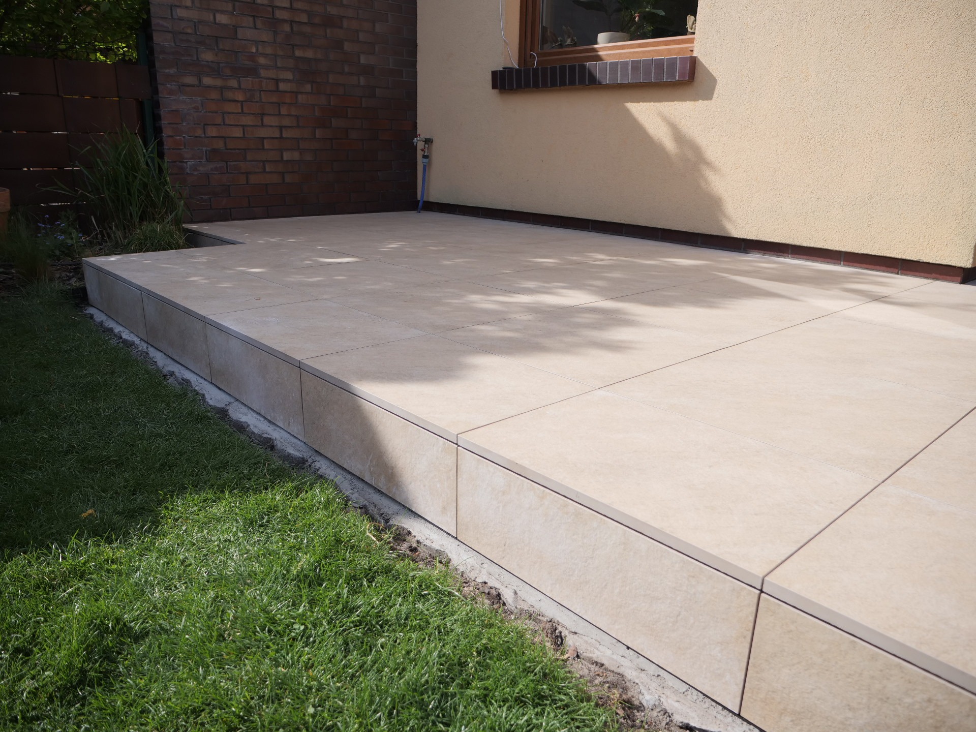 Beżowy taras z płytek ceramicznych przylegający do budynku z brązową cegłą i beżową elewacją, z zielonym trawnikiem w tle. Widok z poziomu gruntu.