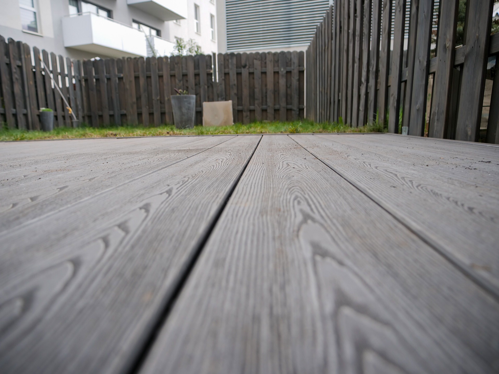 Szary taras z desek kompozytowych, zbliżenie na fakturę drewna. W tle ogrodzenie z ciemnego drewna i fragment nowoczesnego budynku.