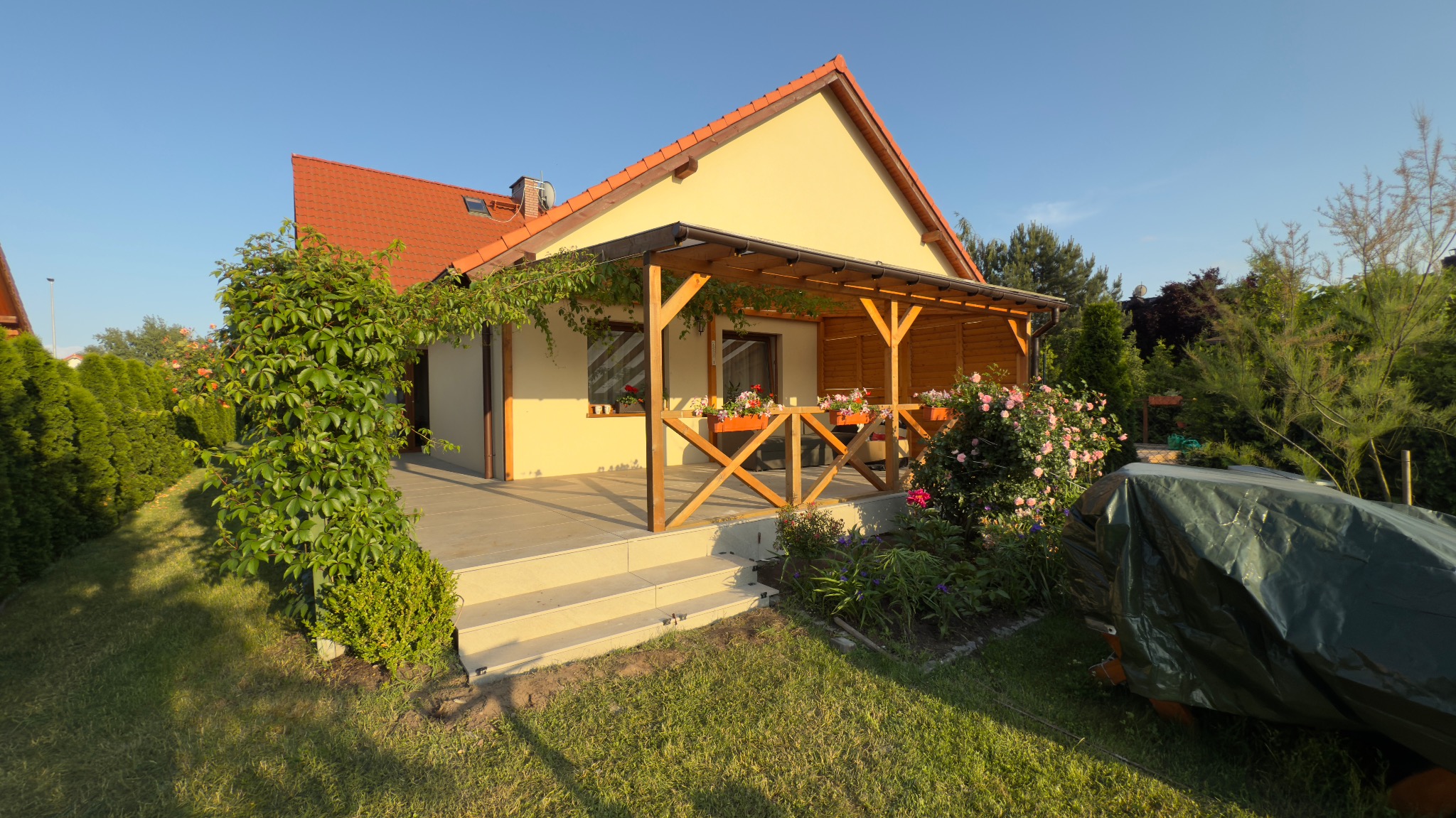 Jasny dom z drewnianą pergolą i tarasem. Kwiaty w skrzynkach na balustradzie. Zieleń pnąca się po ścianie. Zadbany trawnik i roślinność w ogrodzie.