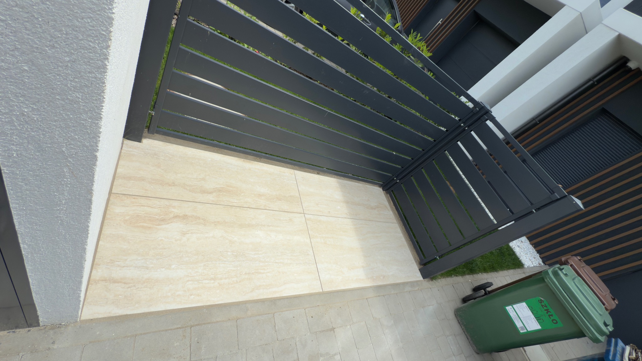 Jasne płytki tarasowe z beżowym wzorem, obok nowoczesne, grafitowe ogrodzenie z poziomych listew. Widoczny fragment elewacji budynku i kosz na śmieci.